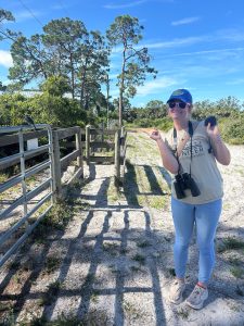 Intern Evelyn in a tan shirt and jeans with binoculars around her neck, pointing to a scrub jay on a nearby fence.