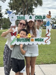 A family of a mother and three children stands holding a life-sized photo frame for take a child outside week.
