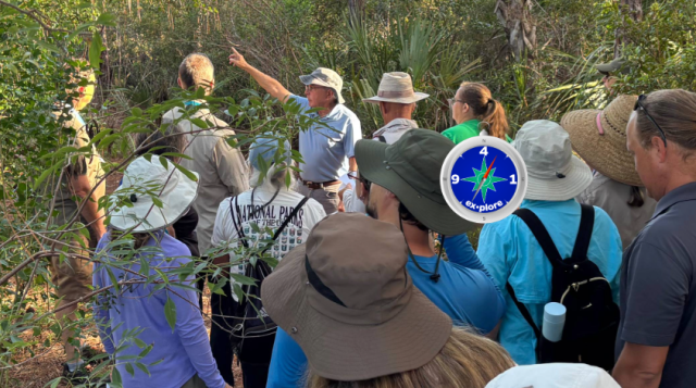 Image of several adults standing in a wooded setting, watching an instructor point to something in the distance