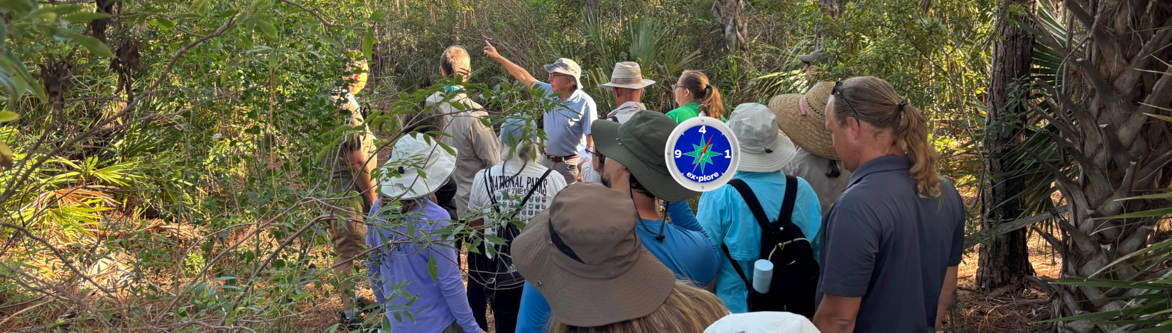 Image of several adults standing in a wooded setting, watching an instructor point to something in the distance