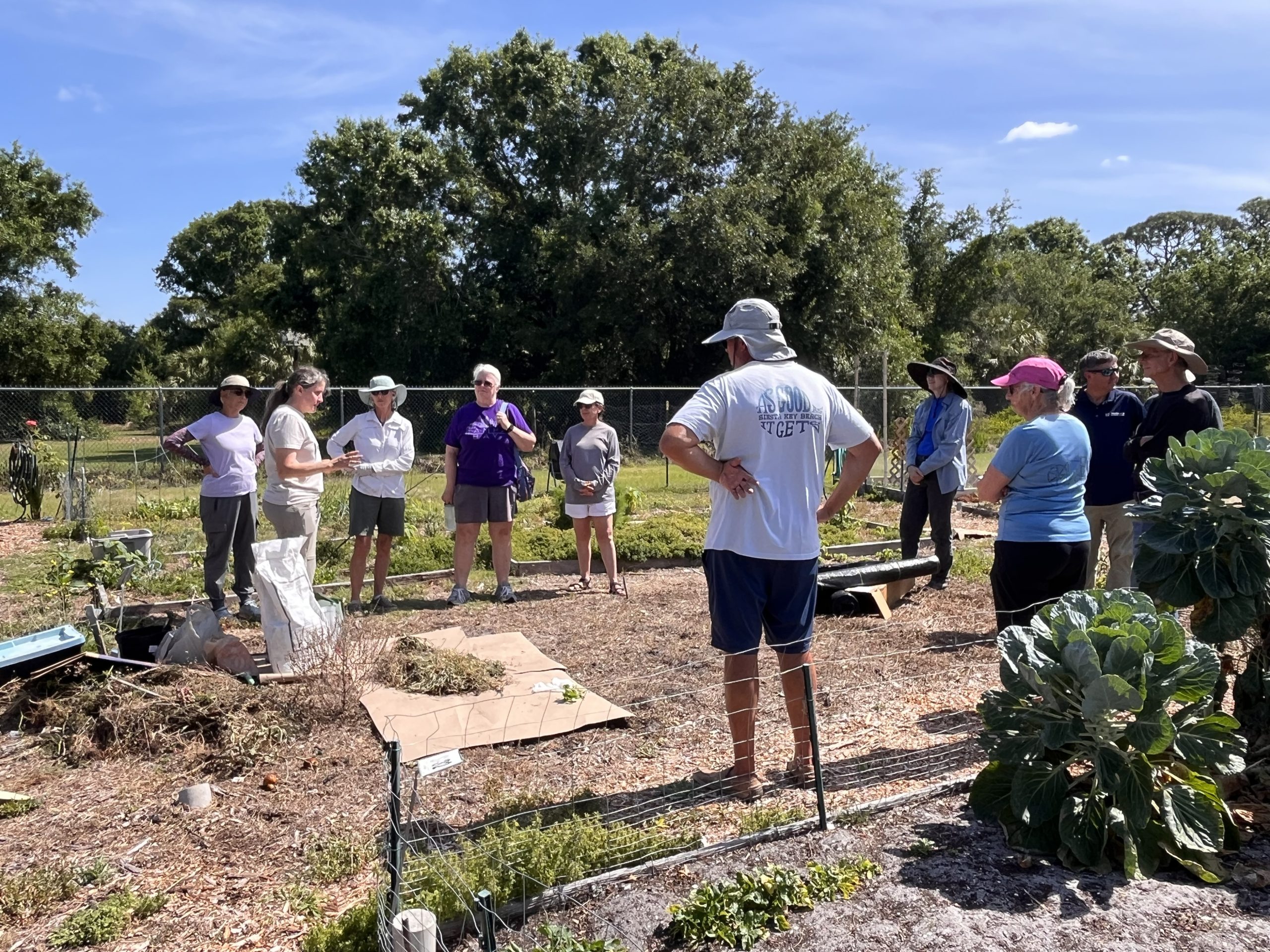 a group of people surrounding a picnic table looking down. Setting is in a community garden thought in this photo we are under the shade of an oak and there are banana fronds in the background along with a fence. On the table are printed resources and we are getting out some seeds to share. 