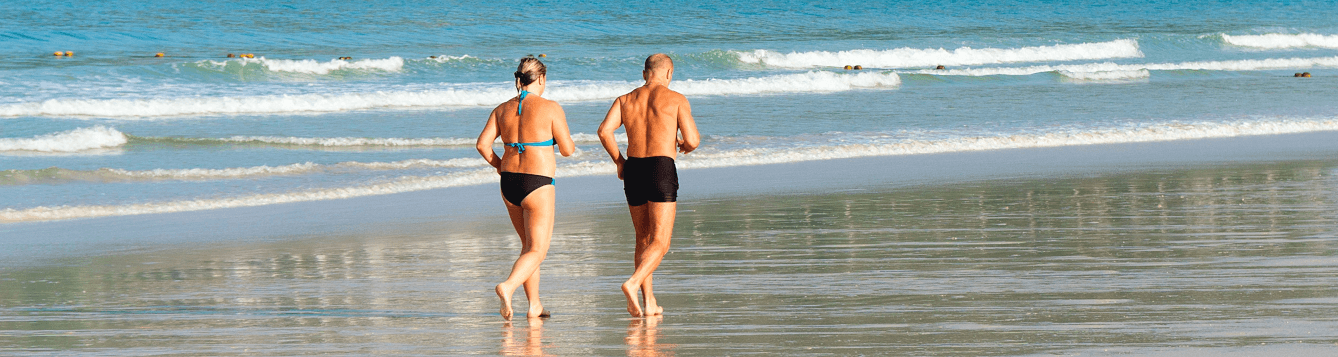 a woman and man jog along a water-soaked beach with gentle waves lapping in background. [credit: pxhere.com]