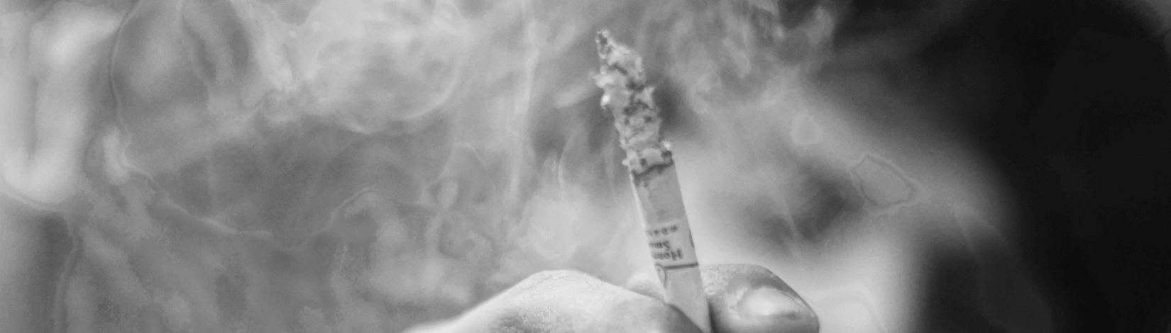 closeup of a cigarette in a hand amidst a haze of smoke, with the smoking man in background. [credit: unsplash.com, arun anoop]