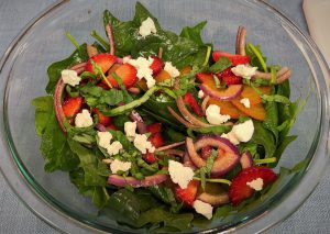 A large glass bowl holds a spinach salad with red onions, strawberries, basil, golden beets, and strawberries. The salad is coated in an oil dressing giving the spinach leaves a shine to them.