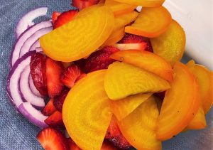 A bowl with purple red onions, red strawberries, and bright orange and yellow golden beets. 