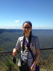 Lydia standing in front of metal chicken wire railing. There is a big blue sky and vast green plains in the background.