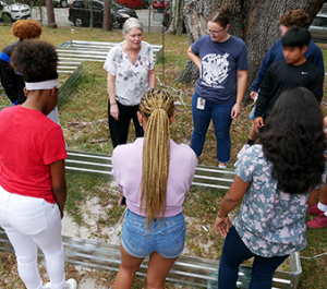 Two women stand by raised garden beds, talking to high school students about sustainability.