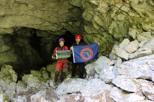 An image of a man and woman standing at the entrance of a cave in Komarnica Canyon. One holds up the National Geographic Society flag and the other holds the Explorers Club Florida Chapter flag.