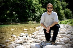 A man in hiking clothes and sunglasses down on one knee by a clear, shallow river in Komarnica Canyon.