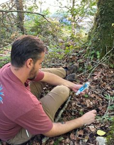 A male scientist sitting on the ground in a wooded area of Komarnica Canyon, measuring soil pH with a sensor.