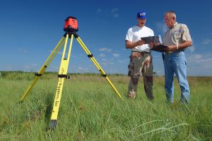 Two men discuss a wetland survey while looking at data on a clipboard. A red surveying tool sits atop a yellow tripod in the foreground.