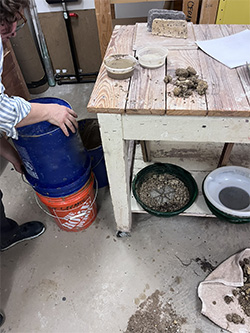A person holds one bucket over another as they begin to sieve clay.