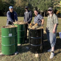 Four people stand by three metal barrels full of straw and wood for firing ceramics.